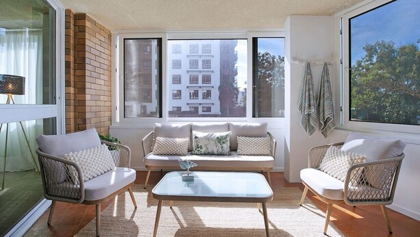 Una sala de estar acogedora con un sofá, dos sillones y una mesa de centro. Amplias ventanas ofrecen vistas a edificios y árboles.