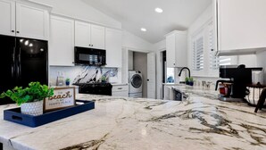 A modern kitchen with white cabinets, a black refrigerator, a microwave, a marble countertop, and a sign that says 'The beach is my happy place'. - The Good Life · Ultimate Gulf Shores Getaway: Hot Tub, RV Pad, Fire-Pit & Beach (Gulf Shores)