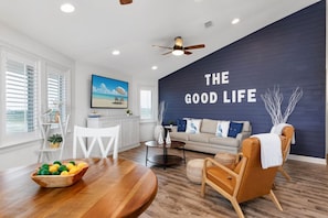 Un soggiorno moderno con una parete decorata in blu scuro, un tavolo da pranzo in legno con una ciotola di frutta, un divano beige e una ventola a soffitto.