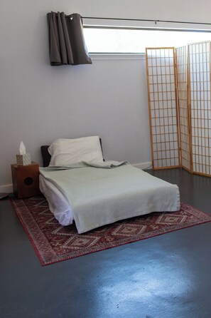 Un dormitorio con una cama, una mesita de noche con una caja de pañuelos, un tapete rojo estampado y un biombos japonés.