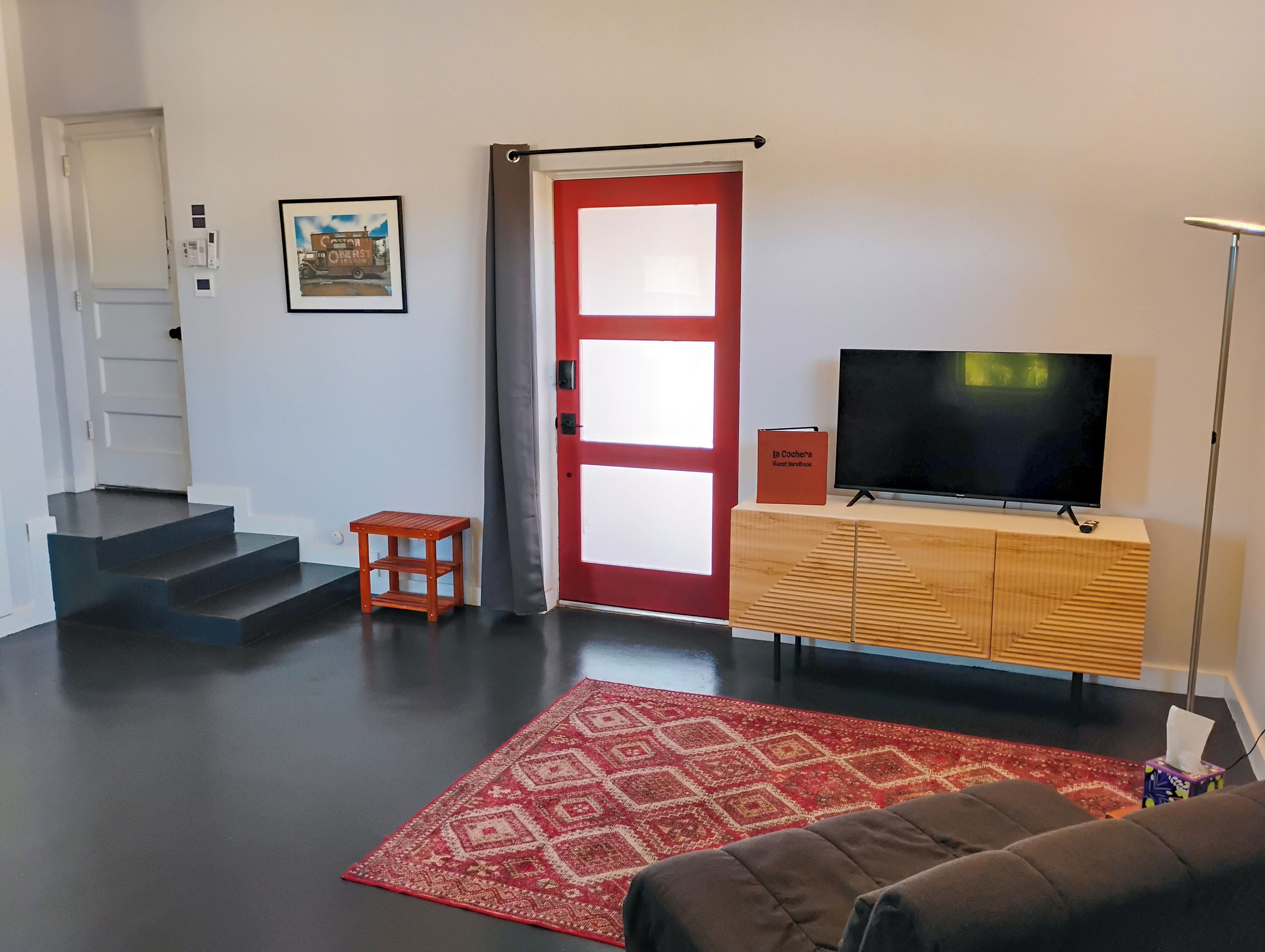 A living room with a TV, a red door, a staircase, and a patterned rug.