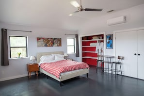 Une chambre à coucher comprenant un lit, des tables de chevet, un ventilateur de plafond, une fenêtre avec des rideaux, un tableau, un meuble de rangement rouge et une petite table avec des chaises.