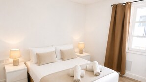 A modern hotel room with a large bed, two bedside tables, a nightstand, a window with curtains, and a ceiling-mounted smoke detector. - Grand Central House - Deluxe Two Bedroom (Gibraltar)