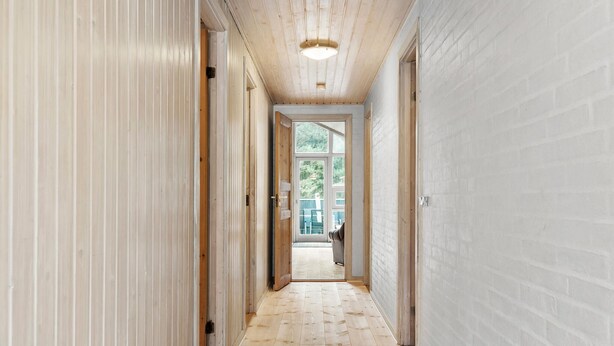 Un pasillo estrecho con pisos de madera y paredes de ladrillo blanco, que conduce a una puerta de vidrio que da a un balcón con mobiliario exterior.