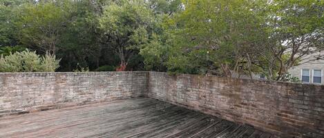 A rooftop deck with a brick wall and wooden flooring, surrounded by trees and a building in the background.