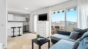 A modern apartment with a kitchenette, a dining area, a living room with a sofa and a TV, and a balcony with a view of the city.
