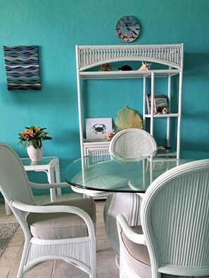 A teal-colored room with a glass-top table surrounded by white wicker chairs. A white shelving unit with decorative items and a clock is mounted on the wall.