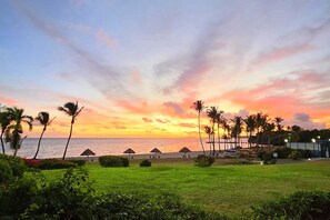 A beach resort with palm trees, thatched-roof huts, and a view of the sea at sunset.