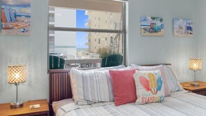 A hotel room with a large window, a bed with striped bedding, two bedside lamps, and wall art depicting beach scenes. - Tropical Isle 215 - Steps to the Beach! (Fort Walton Beach)