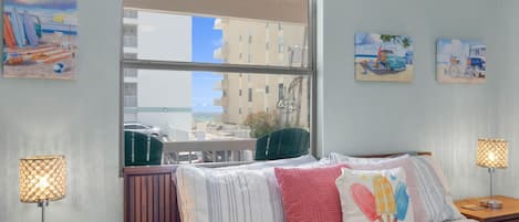 A hotel room with a large window, a bed with striped bedding, two bedside lamps, and wall art depicting beach scenes.