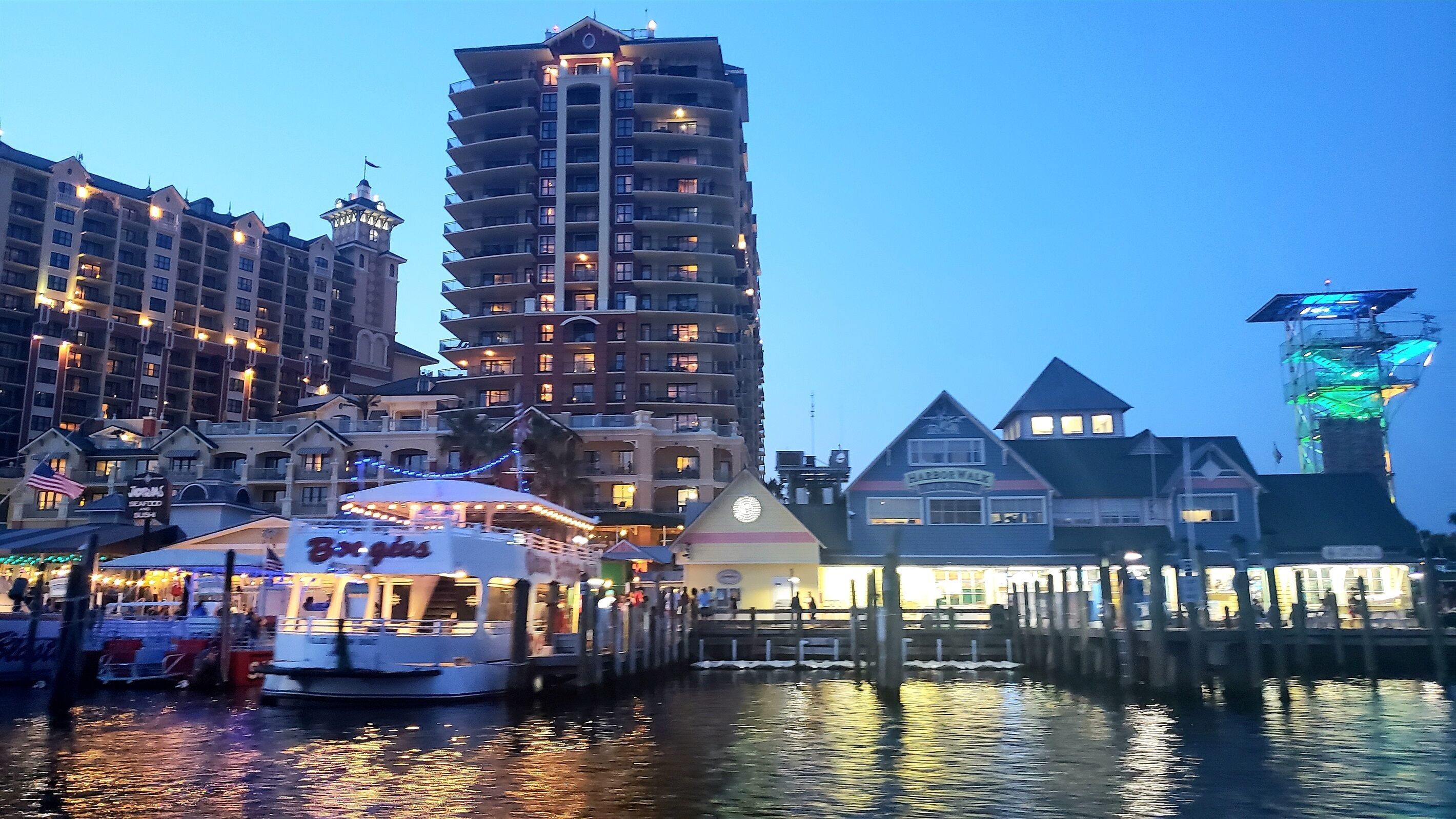 A waterfront area with a tall hotel building, a pier with a boat, and a building with 'Harbor Walk' signage.