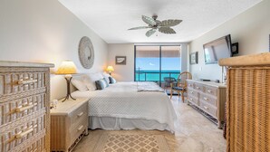 A bedroom with a bed, bedside tables, a ceiling fan, a TV, and a view of the ocean.