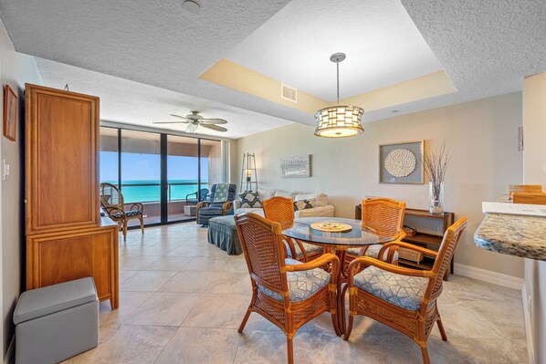 A spacious living area with a large glass door leading to a balcony, wicker furniture, a dining table, and a ceiling fan. - 🩱Luxurious CBC 1-18C: Beach Front Condo | Heated Pool | Gym | Pickleball | Sauna | Hot Tubs! (Clearwater Beach)
