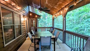 A covered deck with a wooden table and chairs, surrounded by windows and a view of a forest. - Legacy Lodge Cabin-Near Helen,GA Hot Tub,Screened deck, Tubing,Hiking,Loft (Sautee Nacoochee)
