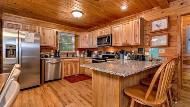 A kitchen with wooden cabinets, a stainless steel refrigerator, a microwave, and a granite countertop island. - Legacy Lodge Cabin-Near Helen,GA Hot Tub,Screened deck, Tubing,Hiking,Loft (Sautee Nacoochee)
