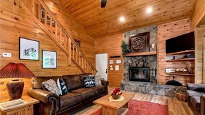 A cozy cabin interior with a wooden staircase, a stone fireplace, a leather sofa, and a television. - Legacy Lodge Cabin-Near Helen,GA Hot Tub,Screened deck, Tubing,Hiking,Loft (Sautee Nacoochee)