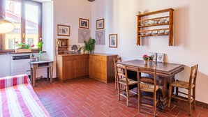 Una cucina accogliente con un tavolo di legno, sedie e un pavimento in mattoni. Alle pareti ci sono quadri appesi e uno scaffale con stoviglie.