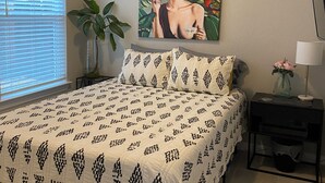 A bedroom with a bed, a nightstand, a lamp, a painting, and a window with blinds. - Frida's Tiny Home (San Antonio)