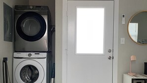 A laundry room with stacked washers and dryers, a door, a small table with a lamp, and a mirror on the wall. - Frida's Tiny Home (San Antonio)
