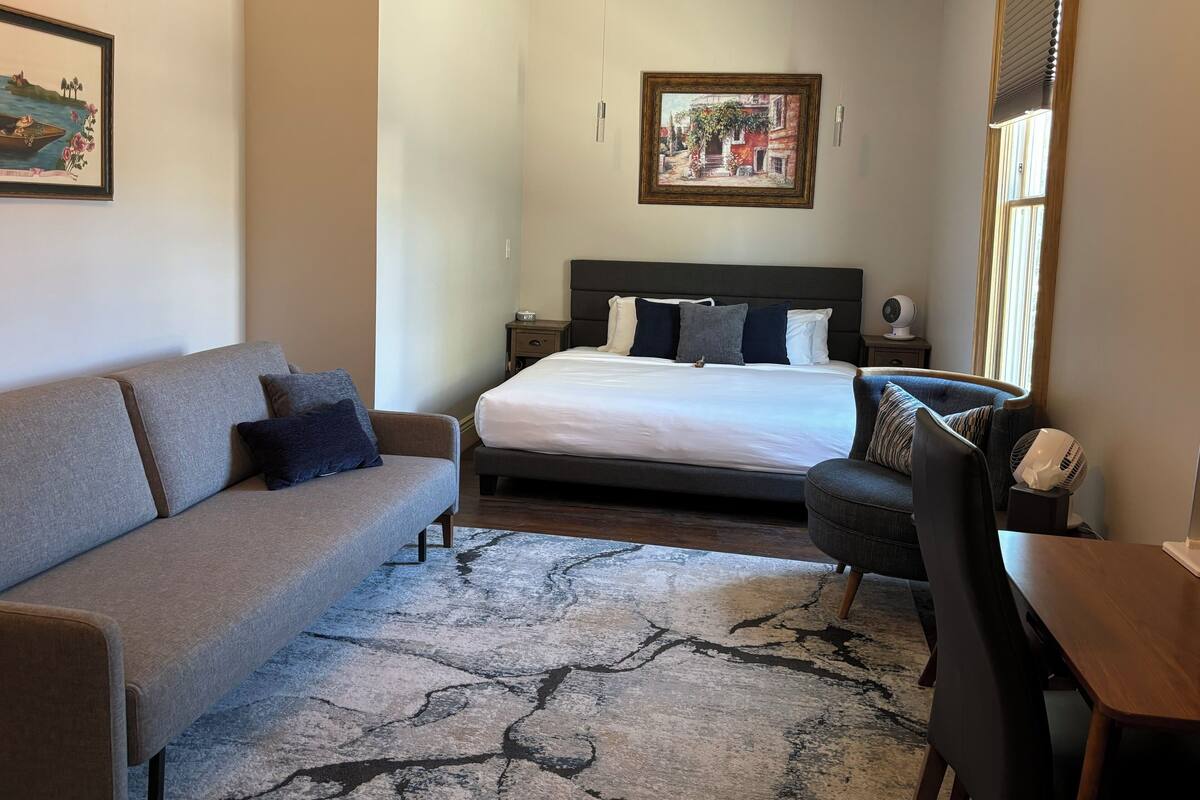 Um quarto com cama, sofá, poltrona, mesa pequena e mesa de cabeceira.