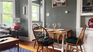 Un coin repas avec une table en bois dressée pour quatre personnes, deux chaises avec des coussins verts, une lampe rouge et un tableau représentant un homard accroché au mur.