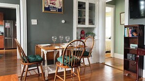 Un coin repas avec une table en bois dressée pour quatre personnes, deux chaises dotées de coussins verts, un tableau encadré représentant des pommes de terre du Maine, et une bibliothèque contenant des livres et une lampe.