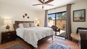 A bedroom with a bed, two bedside lamps, a ceiling fan, a large window with curtains, a small desk, and a chair.