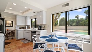 A modern kitchen with white cabinets, a dining area with a round table and chairs, and a large window with a view of greenery.
