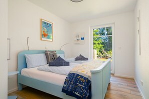 Un dormitorio con cama, una mesita de noche, una lámpara de pared, una ventana con vistas a vegetación y una puerta.