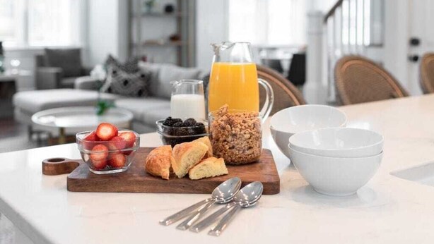 Une table de petit-déjeuner avec un pichet de jus d’orange, un bol de fraises, un bocal de granola et une planche à découper avec des croissants.