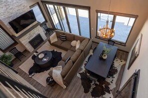 A modern living room with a stone fireplace, a large TV, a sofa, a dining table, and a chandelier.
