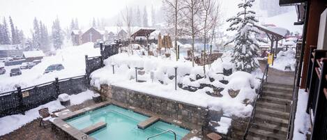 Una piscina con pared de piedra y plataforma de madera, rodeada de suelo y árboles cubiertos de nieve.
