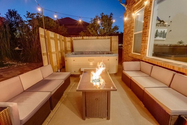 An outdoor patio with a hot tub, cushioned seating, and string lights.