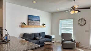 A modern living room with a grey sofa, a wicker ottoman, a ceiling fan, a kitchen island with a granite countertop, and a large window with blinds. - Salty Kisses - 3 miles to beach, pool, gameroom theater room, close to Pier Park (Panama City Beach)