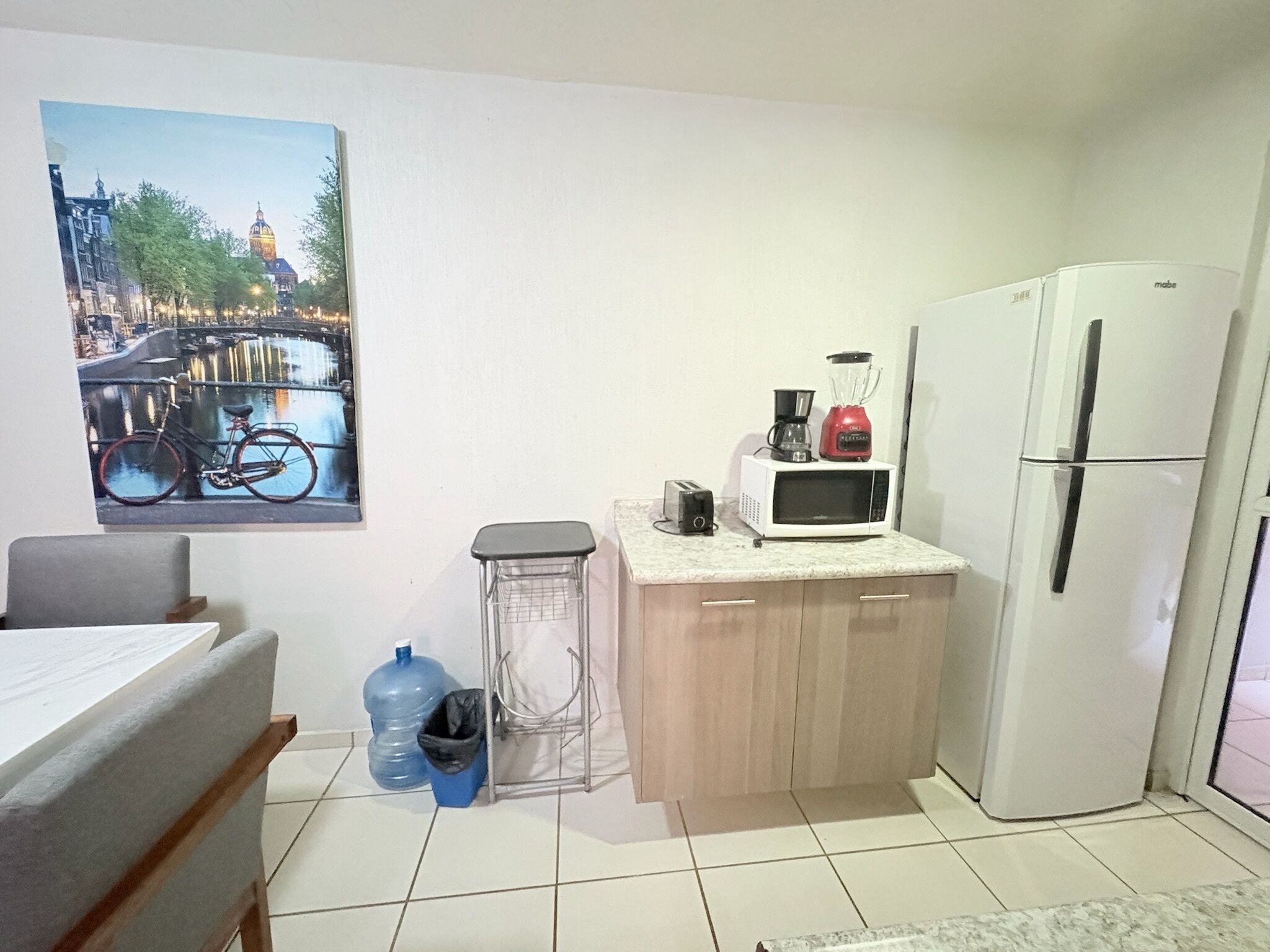 Una pequeña kitchenette con microondas, tostadora y licuadora en la encimera, un refrigerador, un dispensador de agua y un bote de basura.