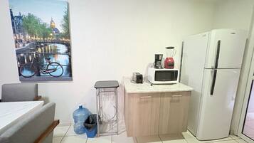 Una pequeña kitchenette con microondas, tostadora y licuadora en la encimera, un refrigerador, un dispensador de agua y un bote de basura.