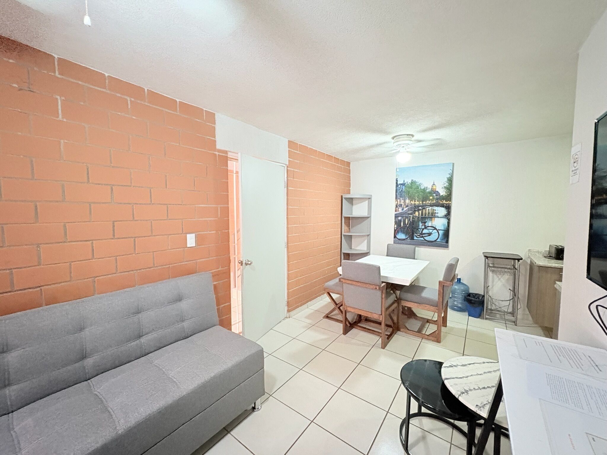 A compact living space with a grey sofa, a dining table with chairs, a small kitchen area, and a door leading to another room.