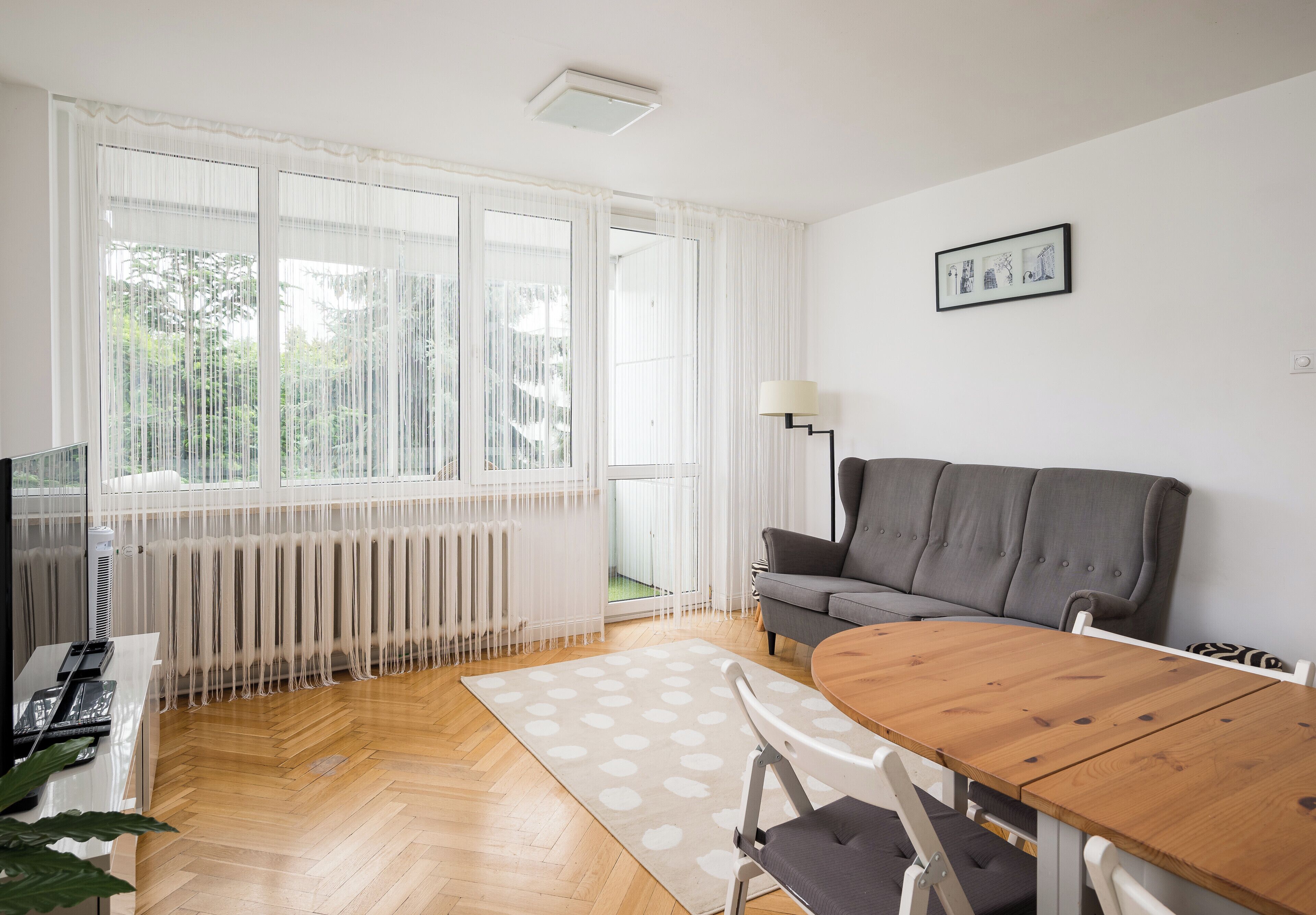 A modern living room with a wooden floor, a grey sofa, a dining table with chairs, a radiator, and a large window with sheer curtains.