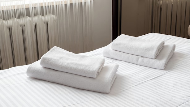 Una cama bien hecha con toallas blancas dobladas encima, un radiador y una ventana con persianas.