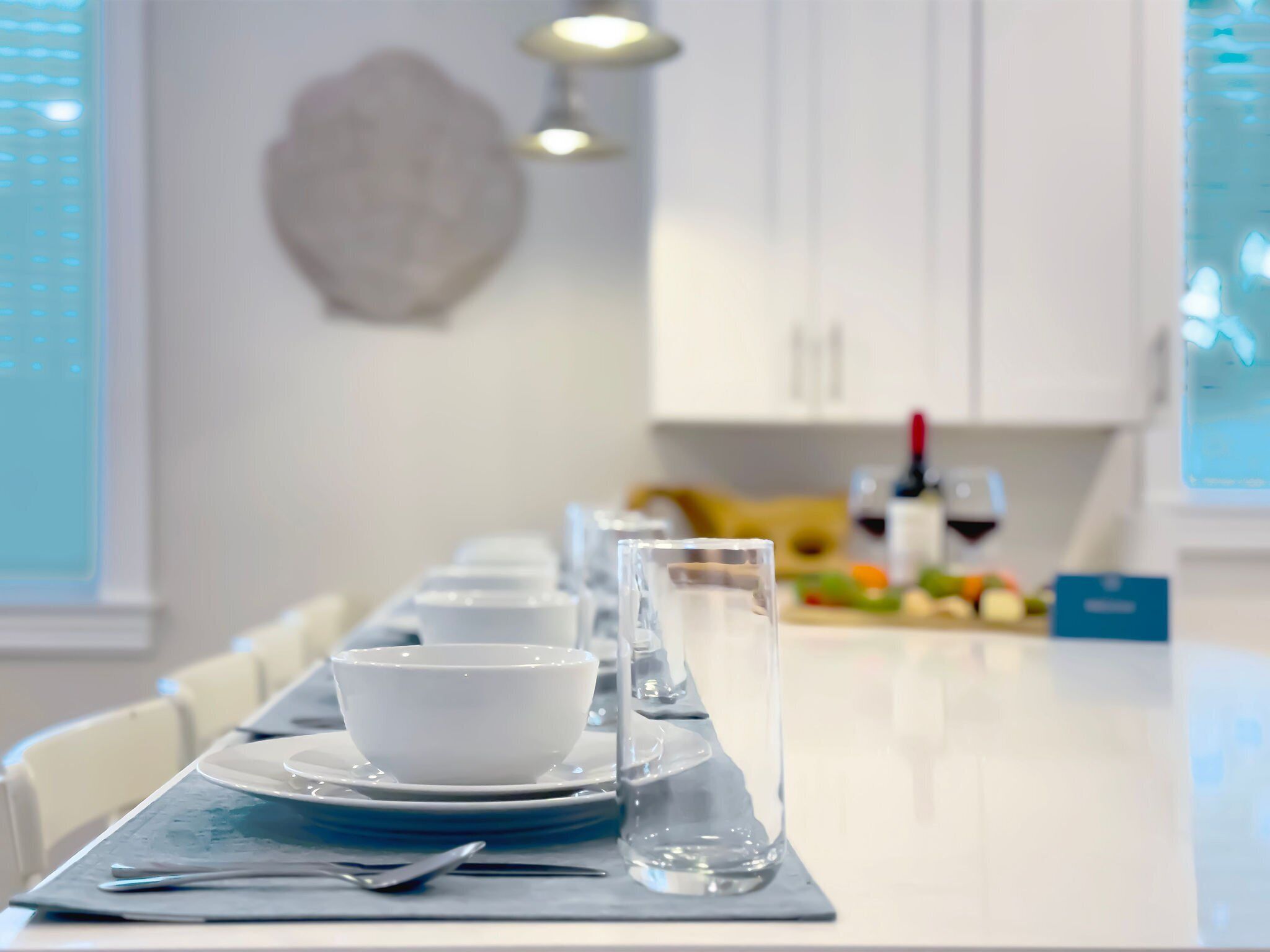 A dining table set with white plates, bowls, and clear glasses, with a modern kitchen in the background.