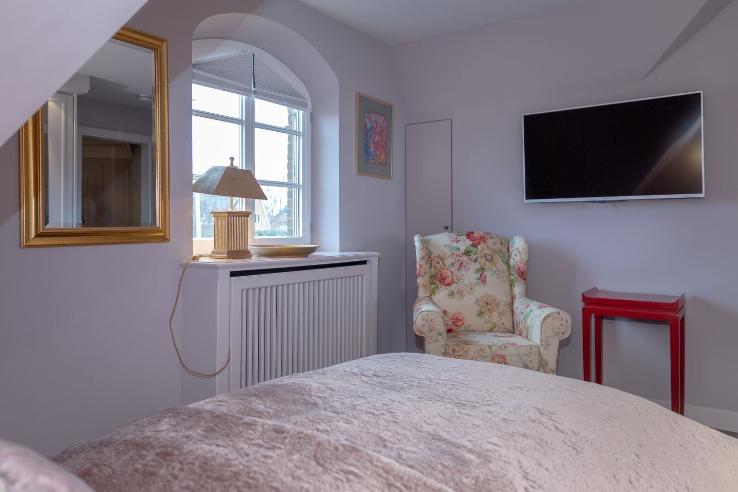 Un dormitorio con cama, radiador, silla, una mesa roja, televisor y ventana con lámpara sobre el radiador.
