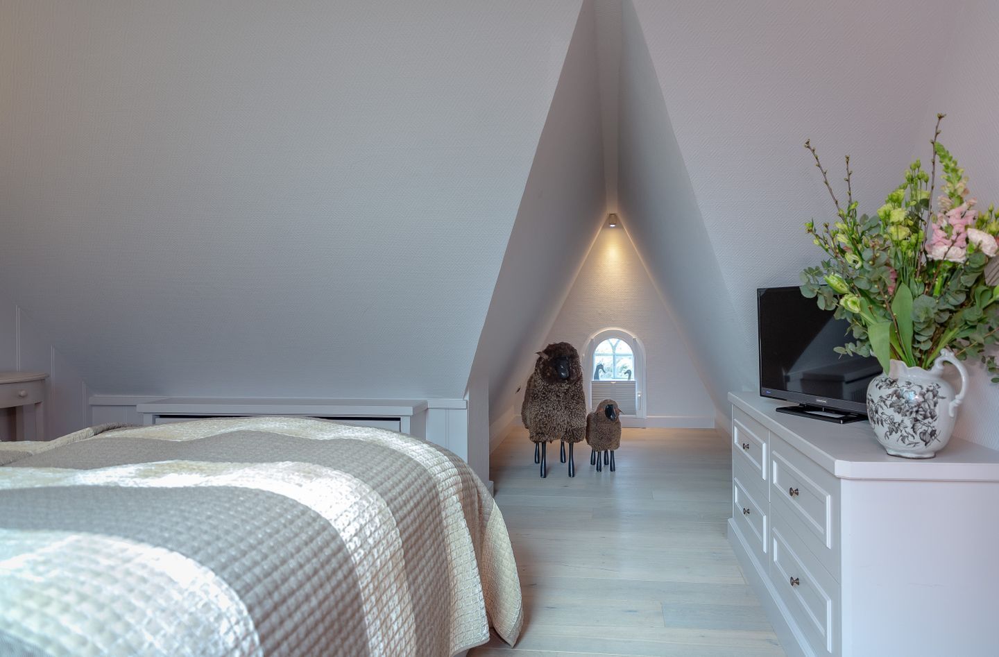 Ein Schlafzimmer mit schrägen Wänden, einem Bett, einer Kommode, einem Fernseher und einem Fenster mit Blick auf Schafe.