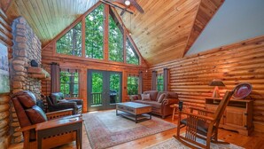 A log cabin interior with a stone fireplace, leather furniture, and large windows offering a view of the forest. - Kind Oak Lake House Peaceful Lakeside Cabin with Hot Tub & Private Dock in Mill Springs (Mill Spring)