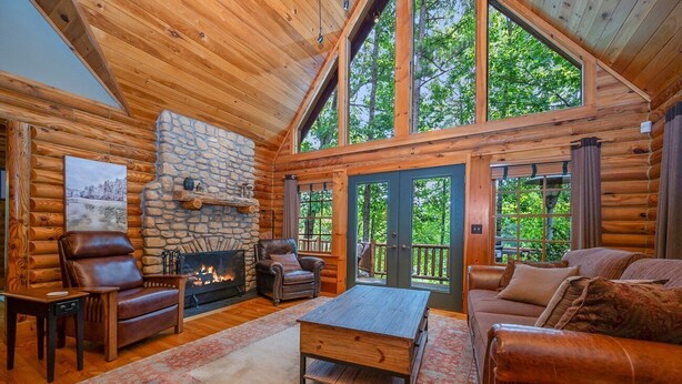 A log cabin interior with a stone fireplace, leather furniture, and large windows offering a view of the forest. - Kind Oak Lake House Peaceful Lakeside Cabin with Hot Tub & Private Dock in Mill Springs (Mill Spring)