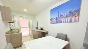 Een kleine keuken met een eethoek, een koelkast, een magnetron en een spoelbak. Op de muur hangt een schilderij van een stadszicht.