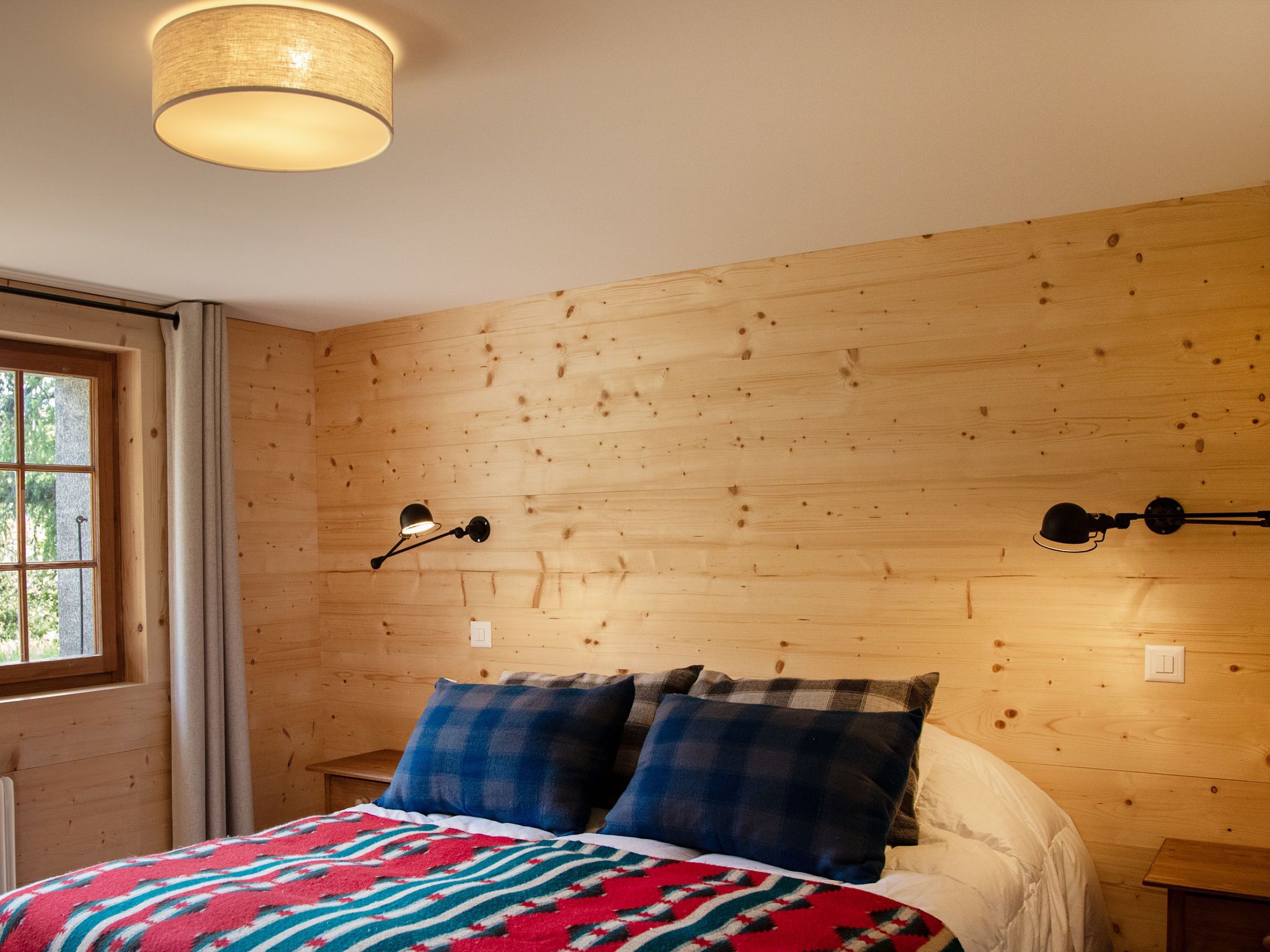A bedroom with a wooden wall, a bed with plaid bedding, a window with curtains, and a wall-mounted lamp.