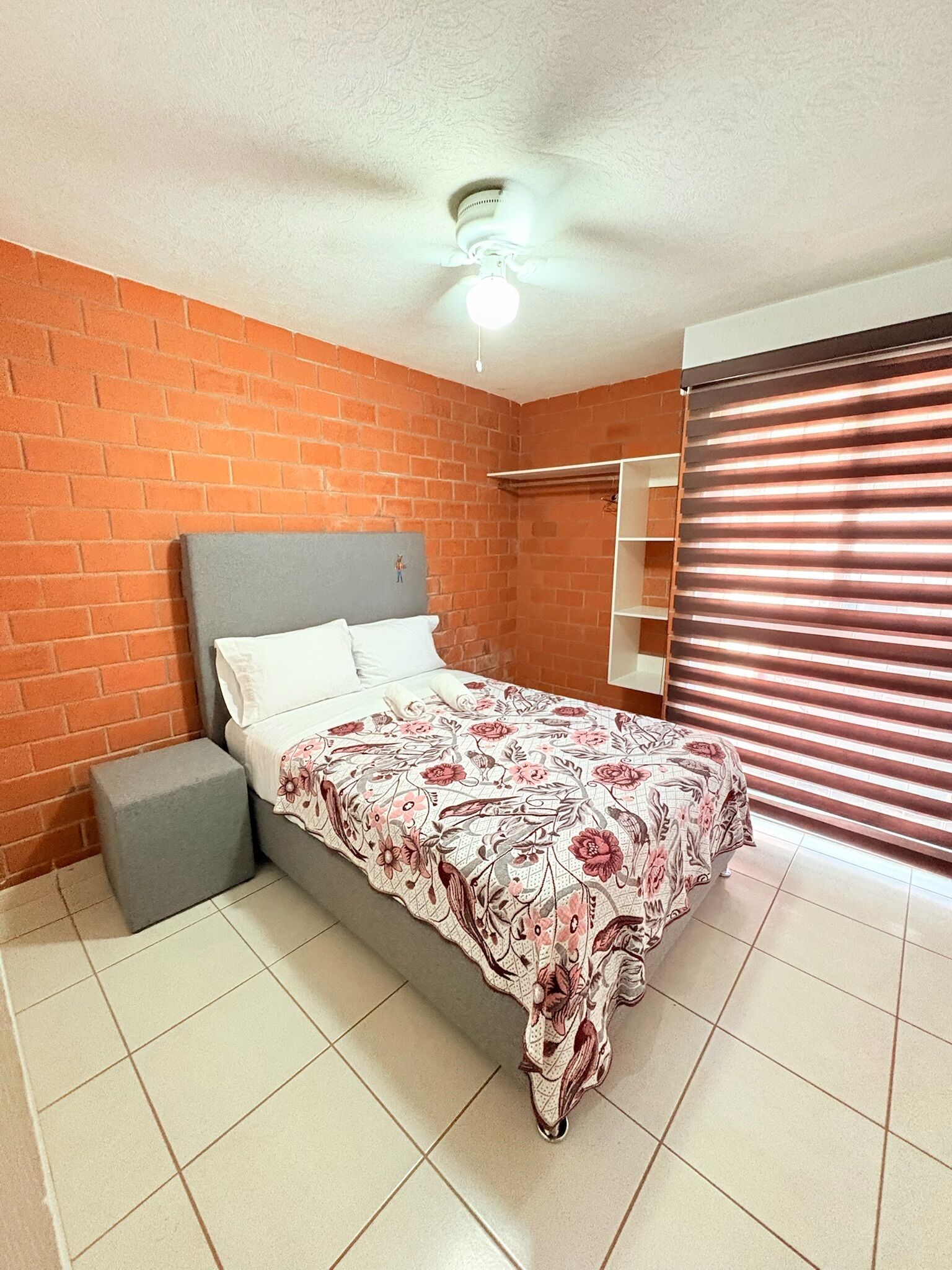 Een slaapkamer met een bakstenen muur, een bed met een bloemenbedekking, een grijze nachtkastje en een plafondventilator.