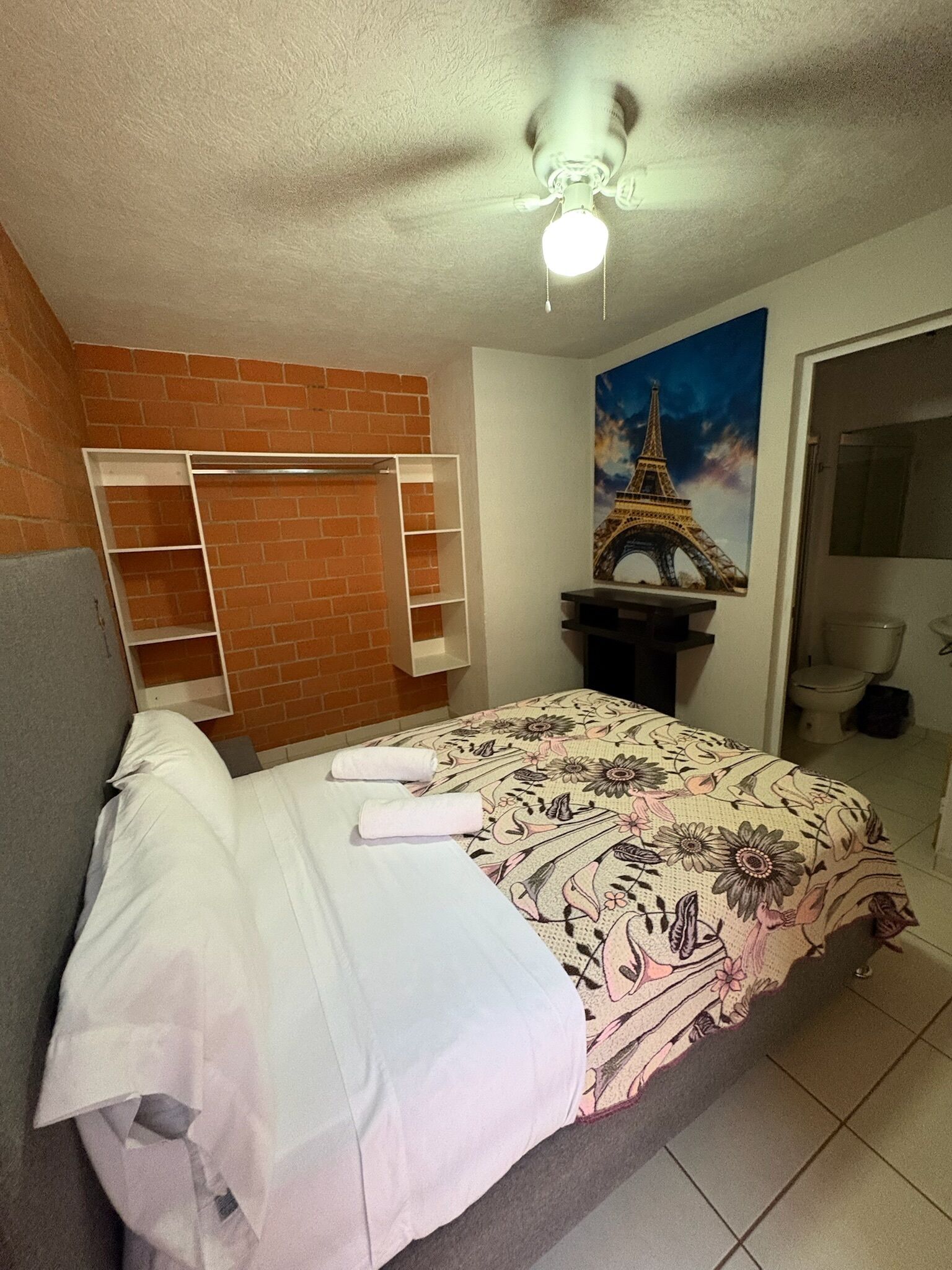 A bedroom with a bed, a ceiling fan, a brick wall, a painting of the Eiffel Tower, and a bathroom visible through an open door.