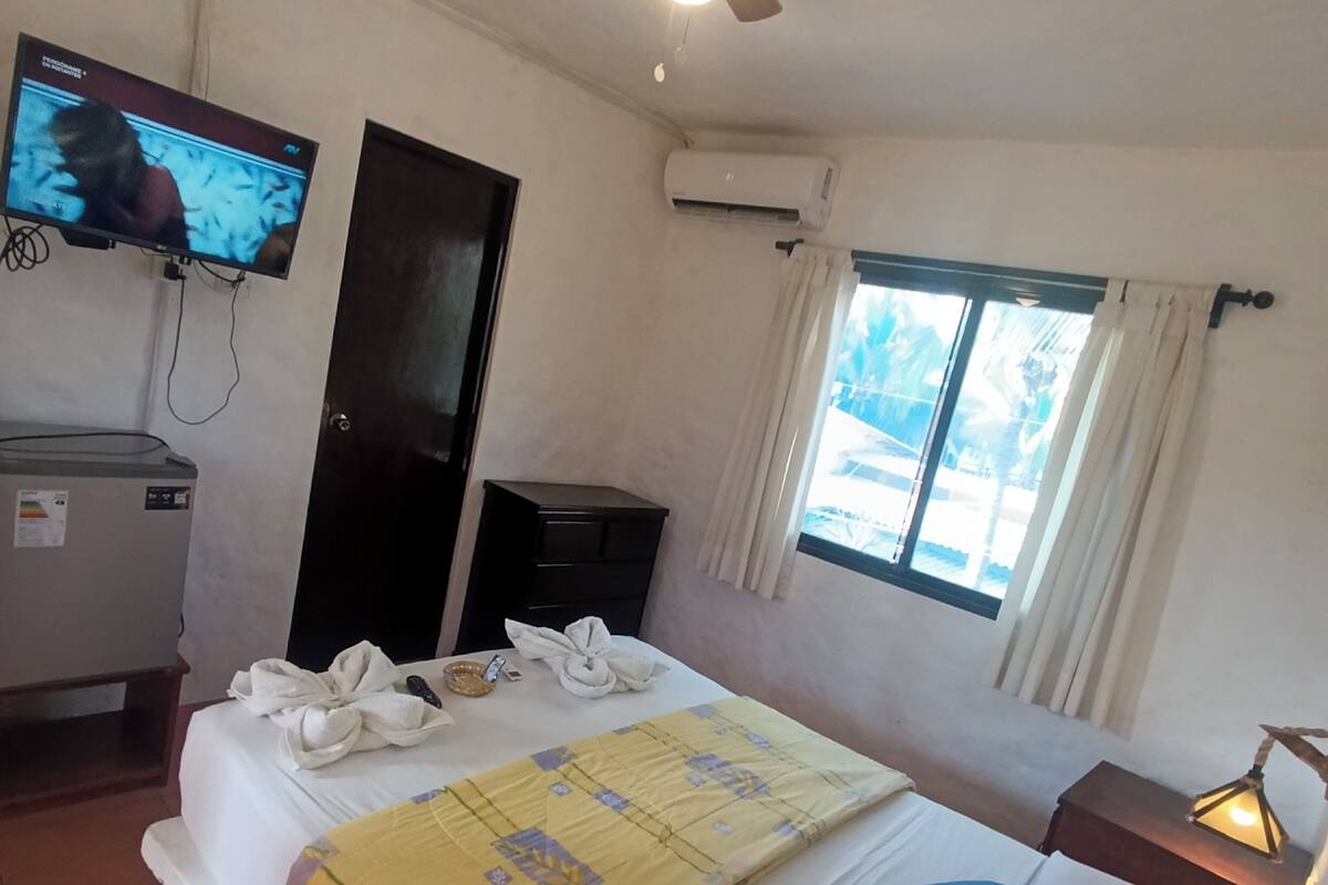 A hotel room with a bed, a TV, an air conditioner, a ceiling fan, a window with curtains, and a bedside lamp.