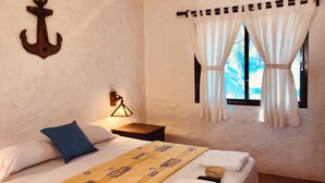 A bedroom with a bed, pillows, a bedside lamp, a window with curtains, and an anchor decoration on the wall. - Aloha Lina (Máncora)